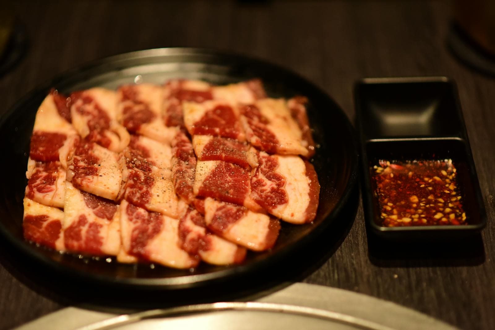 Các miếng 갈비살 thịt bò 한우 có vân mỡ trên đĩa đen tại nhà hàng nướng Hàn Quốc, kèm nước chấm ớt