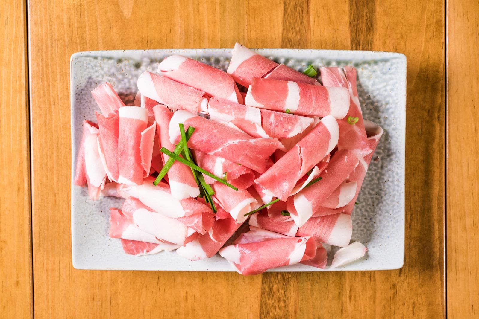 Các lát 우삼겹 thịt bụng bò thái mỏng cuộn tròn với sọc mỡ trắng, bày cùng hành lá