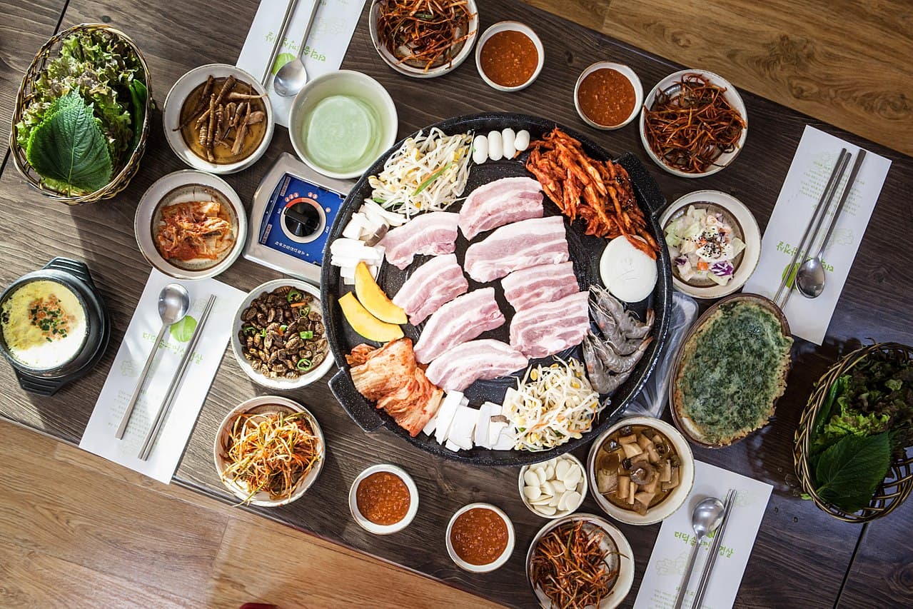 Nhìn từ trên xuống một bàn nướng thịt Hàn Quốc với các lát 삼겹살 trên vỉ nướng tròn, xung quanh là các đĩa banchan, rau cuộn và nước chấm