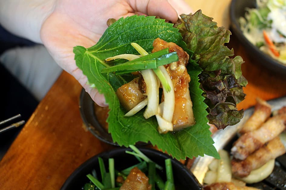 Một cuộn 쌈 được tạo trong lòng bàn tay mở: rau diếp đỏ đặt lá tía tô, một miếng thịt ba chỉ nướng, các lát tỏi, hành lá và chút 쌈장