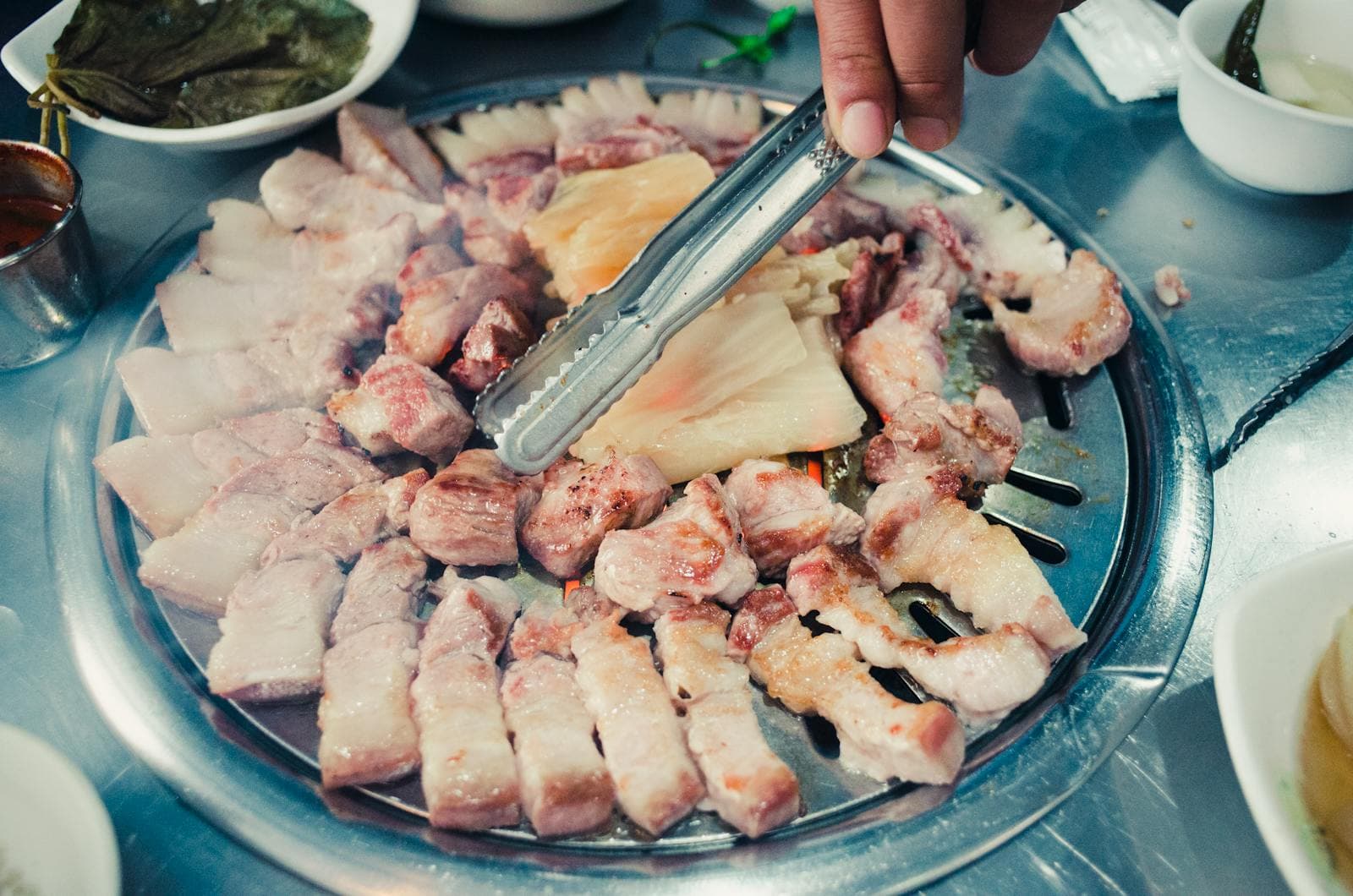 Một bàn tay đang dùng kẹp inox để lật các lát 삼겹살 trên vỉ nướng tròn